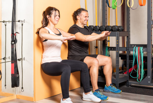 Du žmonės sporto salėje išlaiko pritūpimo padėtį prie sienos