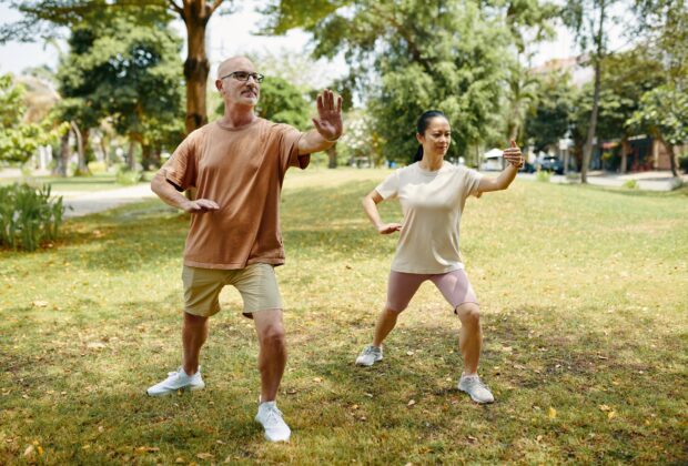 Du žmonės parke atlieka tai chi judesius, iškėlę rankas ir perkeldami kūno svorį.